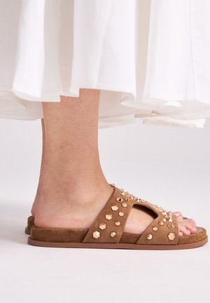 Pies con sandalias tipo slide de ante marrón con tachuelas geométricas doradas, combinadas con una falda o vestido blanco y fluido.