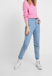 Hellblaue, hochgeschnittene Jeans kombiniert mit einem rosa strukturierten Strickpullover und weißen High-Top-Sneakern. Fokus auf lässige, lockere Passform.