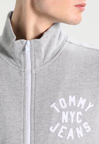 Grijze ritstrui van zacht materiaal, met een hoge kraag en een witte logo-opdruk met de tekst "TOMMY NYC JEANS" op de borst.