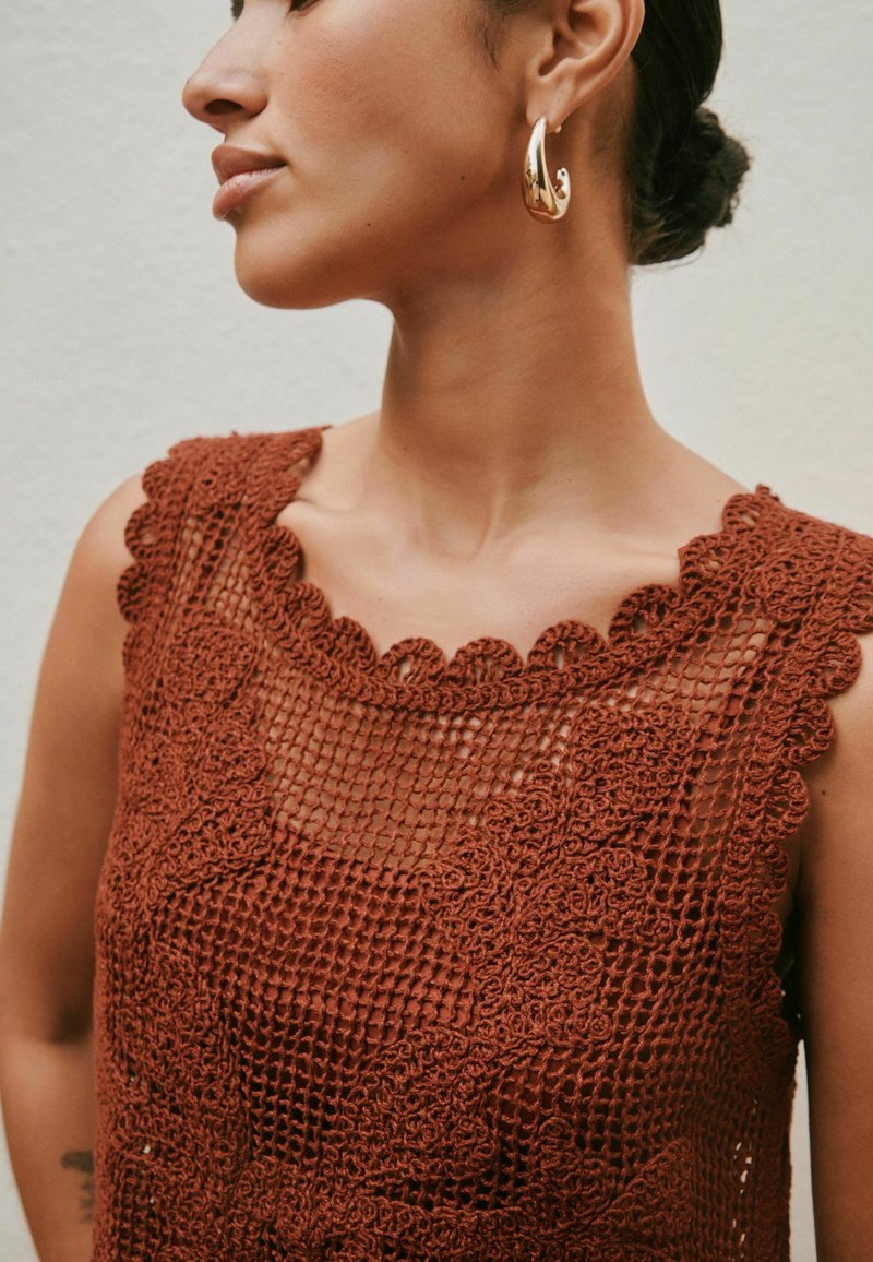 Femme portant un haut sans manches en crochet couleur rouille et des boucles d'oreilles créoles dorées, les cheveux attachés en chignon, regardant vers la gauche.