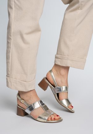 Voeten met metallic zilveren sandalen met open teen en blokhak, gecombineerd met beige opgerolde ribfluwelen broek op een lichte achtergrond.