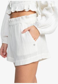 Shorts blancs texturés avec une taille élastique et des poches latérales. Présente un détail en bouton argenté sur la poche. Coupe décontractée et ample.