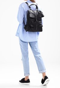 Černý kožený batoh s nastavitelnými popruhy, přední klopou a dvěma vnějšími kapsami. Nosený s modrou pruhovanou košilí a světlými džíny.