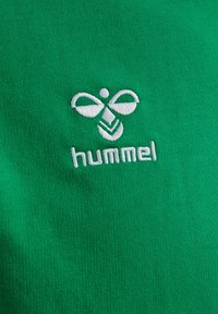 White embroidered hummingbird logo and "hummel" text on green fabric background, representing sportswear brand emblem.