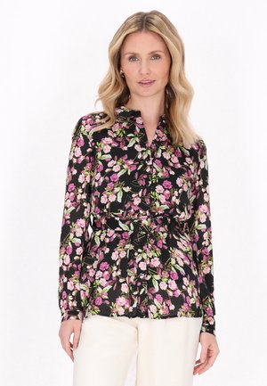 Frau trägt eine schwarze Bluse mit rosa und grünem Blumenmuster, langen Ärmeln und weißen Hosen, vor weißem Hintergrund stehend.