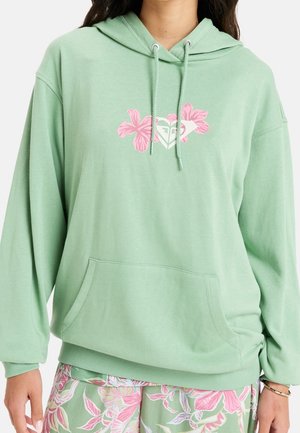Jasnozielona bluza z kapturem z kieszenią z przodu i sznurkami, z różowym kwiatowym logo na piersi, noszona z dopasowanymi kwiatowymi spodniami.