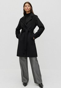 Svart ullkappa med bälten i midjan, dubbelknäppt design, slagkrage och sidofickor, kombinerat med grå vida byxor.