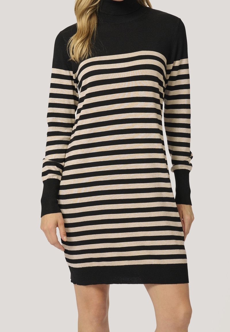 Femme portant une robe-pull à col roulé à manches longues rayée horizontalement en noir et beige, posant devant un fond uni.
