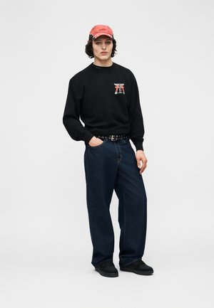 Νεαρός άνδρας που φορά κόκκινο καπέλο, μαύρο φούτερ με γραφικό, σκούρα τζιν με φαρδιά πόδια, μαύρη ζώνη με τρουκς και μαύρα αθλητικά παπούτσια, στέκεται με το ένα χέρι στην τσέπη.