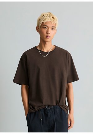 Junge Person mit kurzen blonden Haaren, die ein lockeres braunes T-Shirt, dunkle Hosen und eine silberne Kettenhalskette vor grauem Hintergrund trägt.