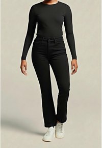 Personne portant un haut noir à manches longues, un pantalon noir taille haute et des baskets blanches, debout devant un fond uni.