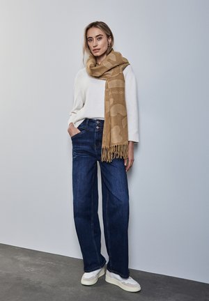 Sciarpa beige a fantasia con frange, maglione bianco, jeans a gamba larga blu scuro e sneakers bianche. Sfondo neutro, texture morbide.