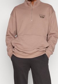 Hnědá mikina na zip s vysokým límcem, na ní je logo Reebok. Materiál vypadá měkký a texturovaný, doplněný tmavě šedými kalhotami.