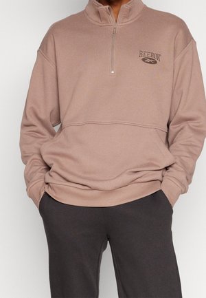 Brauner Reißverschluss-Sweatshirt mit hohem Kragen, versehen mit einem Reebok-Logo. Der Stoff wirkt weich und strukturiert, kombiniert mit dunkelgrauen Hosen.