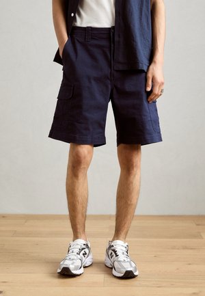 Homme portant un short cargo bleu marine, un t-shirt blanc, une chemise bleu marine ouverte et des baskets blanches, debout sur un sol en bois.