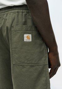 Κοντινό ενός χεριού που ακουμπά δίπλα σε ελιάς πράσινο σορτς Carhartt με ελαστική μέση και πίσω τσέπη με το λογότυπο της μάρκας.