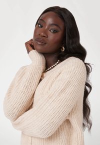 Hellbeige gestrickter Pullover mit geripptem Muster, langen Ärmeln und einem leicht übergroßen Schnitt, kombiniert mit einer Perlenkette und Creolen.