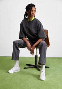 Šedá oversized mikina, černé džíny a bílé tenisky. Zelené tričko viditelné na límci. Pletený účes. Minimalistické prostředí.