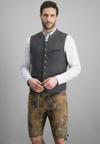 Graham Wolle Weste mit Metallknöpfen über einem weißen Hemd, kombiniert mit beigen Lederhorts mit blauer Stickerei und seitlichen Bindebändern.