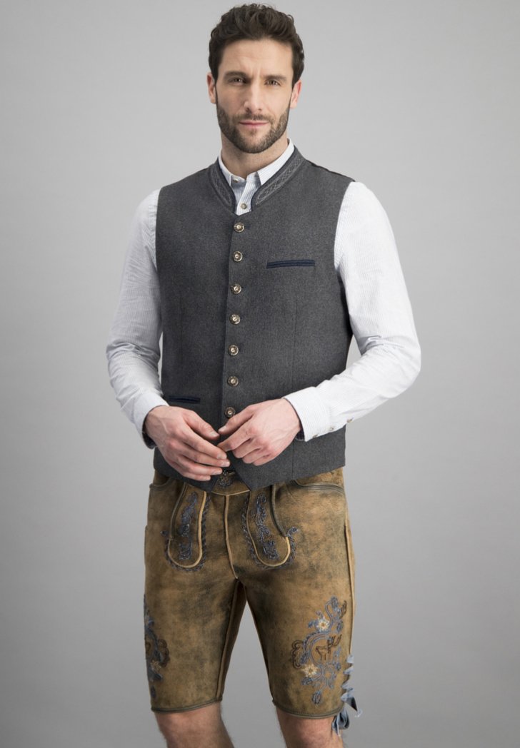 Graham Wolle Weste mit Metallknöpfen über einem weißen Hemd, kombiniert mit beigen Lederhorts mit blauer Stickerei und seitlichen Bindebändern.