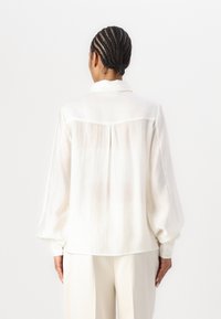 Weiße, leichte Bluse mit langen Puffärmeln, sanfter Textur, Kragen und einem rückenliegenden Yoke-Nähten. Runden Sie das Ganze mit einer lässigen Passform ab.