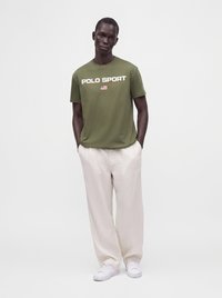 Άνδρας που φοράει μπλούζα Polo Sport σε ελαιί πράσινο, μπεζ παντελόνια και λευκά αθλητικά παπούτσια, στέκεται με τα χέρια στις τσέπες, μπροστά από ένα μονόχρωμο φόντο.