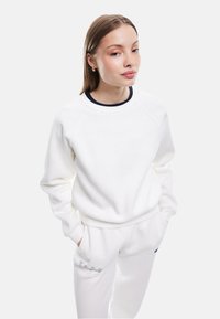 Sudadera blanca con cuello de canalé marino, mangas raglán y corte holgado. Combinada con pantalones deportivos blancos con texto bordado.