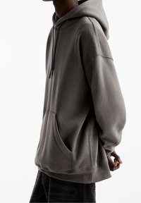 En person klädd i en överdimensionerad grå huvtröja med ficka framtill och svarta dragsnören, står mot en vit bakgrund.