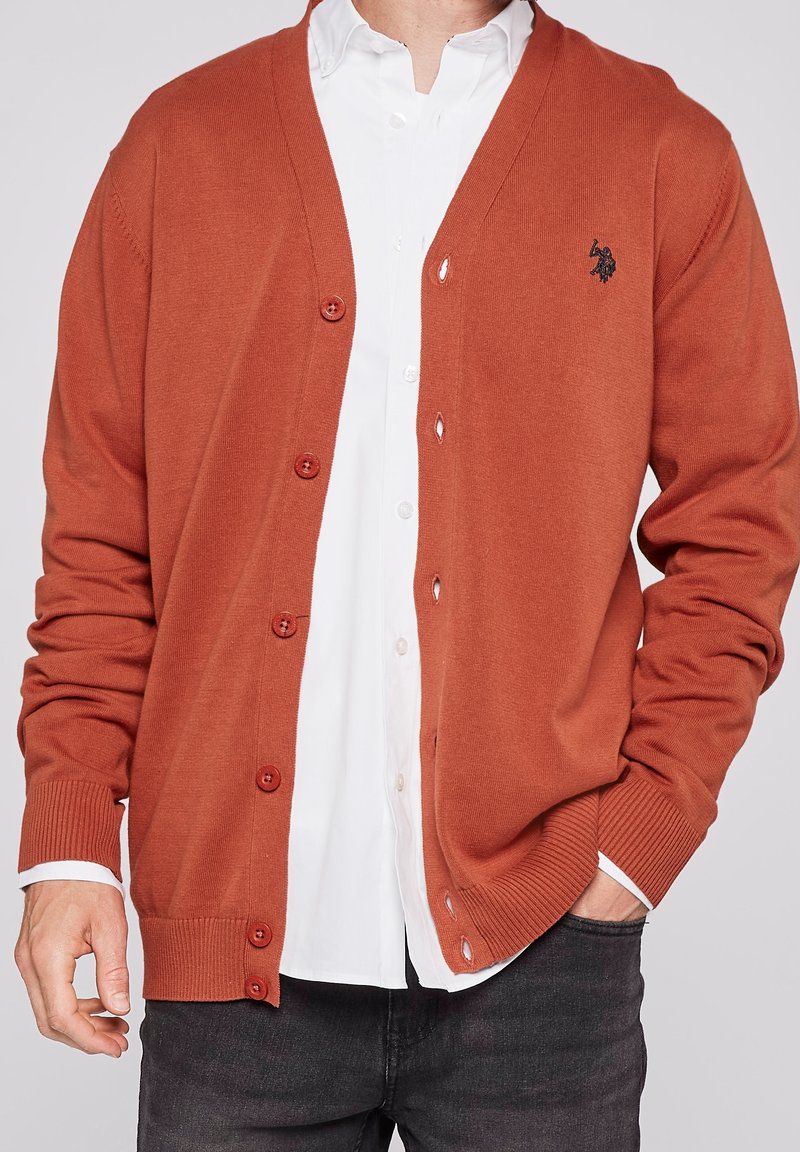 Rust-colored cardigan made of knit material, featuring a V-neck, front buttons, and ribbed cuffs, layered over a white shirt.