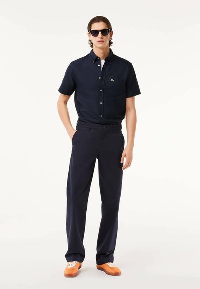 Chemise navy à manches courtes avec col, sous un t-shirt blanc, associée à un pantalon navy foncé et des baskets orange. Des lunettes de soleil noires sont portées.