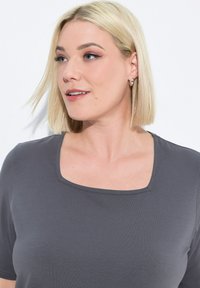 Grijze top met korte mouwen en een vierkante halslijn, gemaakt van gladde stof met een subtiele textuur. Het model heeft steil blond haar.
