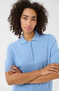 Lichtblauw polo shirt, korte mouwen, zachte textuur, drieknoopsluiting, ribgebreide kraag en manchetten. Model met gekruiste armen op een neutrale achtergrond.
