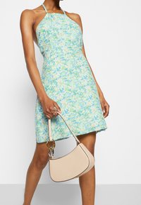 Blommig halterneck-klänning i ljusblå och gröna nyanser, figurnära midja, utsvängd kjol, matchad med en beige handväska med guldaccent.