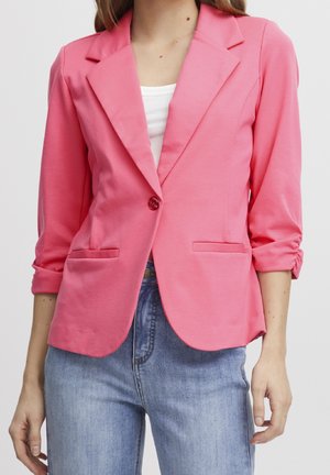 Kobieta ubrana w jaskraworóżową marynarkę na jeden guzik z marszczonymi rękawami 3/4, na białej bluzce i niebieskich dżinsach, stojąca na neutralnym tle.