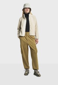 Chaqueta de forro polar beige, cuello alto negro, pantalones cargo verdes y gorra tipo cubo gris. Materiales texturizados y bolsillos visibles en los pantalones. Zapatillas.