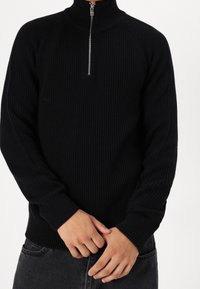 Pull-over noir côtelé avec un col montant et une demi-fermeture éclair à l'avant. Comprend des manches longues et un tissu en maille texturée pour la chaleur.