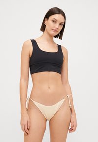 Juodas supjaustytas bikini viršus su kvadratine iškirpte, derinamas su šviesiai smėliniais nerti stiliaus bikini apatiniais, turinčiais šonines juostas ir karoliukų akcentus.