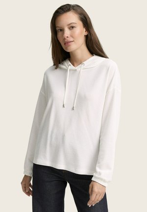 Femme aux longs cheveux bruns portant un sweat à capuche blanc à rayures et un jean sombre, se tenant devant un fond clair uni.