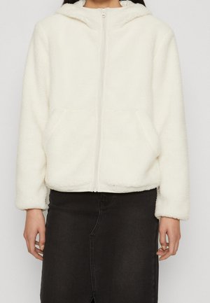 Personne portant un sweat à capuche en polaire blanche avec fermeture éclair et poches, et une jupe noire, se tenant devant un fond clair uni.