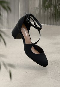 Scarpa nera con tacco a rotondo, tomaia in tessuto morbido, cinturino alla caviglia e tacco a blocco texturizzato, esposta su uno sfondo neutro.