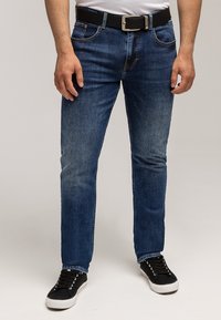 Mężczyzna ubrany w obcisłe niebieskie dżinsy, czarny pasek, białą koszulkę i czarne trampki, stojący na jednolitym jasnym tle.