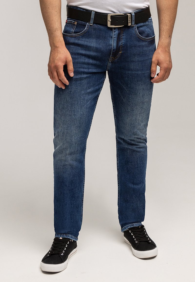 Mężczyzna ubrany w obcisłe niebieskie dżinsy, czarny pasek, białą koszulkę i czarne trampki, stojący na jednolitym jasnym tle.