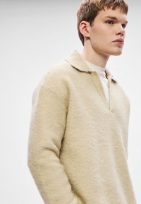 Béžový mikina z fleecu s výstrihom do V a golierom, so štruktúrovaným povrchom a spustenými ramenami pre pohodlný strih.