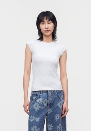 Jeune femme aux cheveux noirs portant un top blanc ajusté sans manches et un jean bleu ample avec un léger motif floral sur un fond uni.