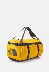 Yellow duffel bag with black straps and accents. Features logo on the side, durable fabric, and adjustable carrying strap. Rounded shape.