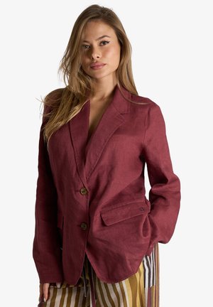 Donna dai capelli lunghi che indossa un blazer bordeaux abbottonato e pantaloni a righe multicolore, posando con una mano in tasca su sfondo bianco.