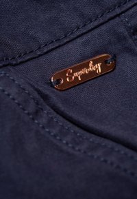 Navy tyg närbild med en liten, bronzfärgad Superdry-logotag och kontrasterande sömmar. Strukturerad yta med synliga sömmar.