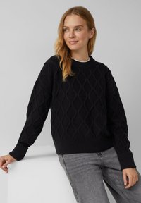 Czarny sweter dzianinowy z teksturowanym wzorem diamentowym, okrągłym dekoltem oraz ściągaczami przy rękawach i u dołu. W połączeniu z szarymi dżinsami.
