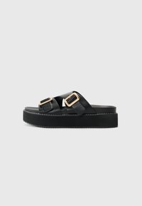 Sandales plateformes noires avec deux brides croisées, ornées de boucles en ton or. En cuir lisse et semelle extérieure texturée.