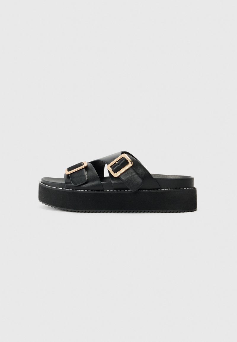 Sandales plateformes noires avec deux brides croisées, ornées de boucles en ton or. En cuir lisse et semelle extérieure texturée.